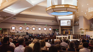 A congregation inside a church, seated and standing, with a screen displaying "Twentieth Sunday in Ordinary Time." Warm lighting and organ pipes seen.