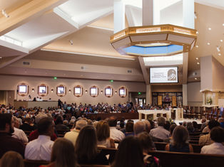 A congregation inside a church, seated and standing, with a screen displaying "Twentieth Sunday in Ordinary Time." Warm lighting and organ pipes seen.