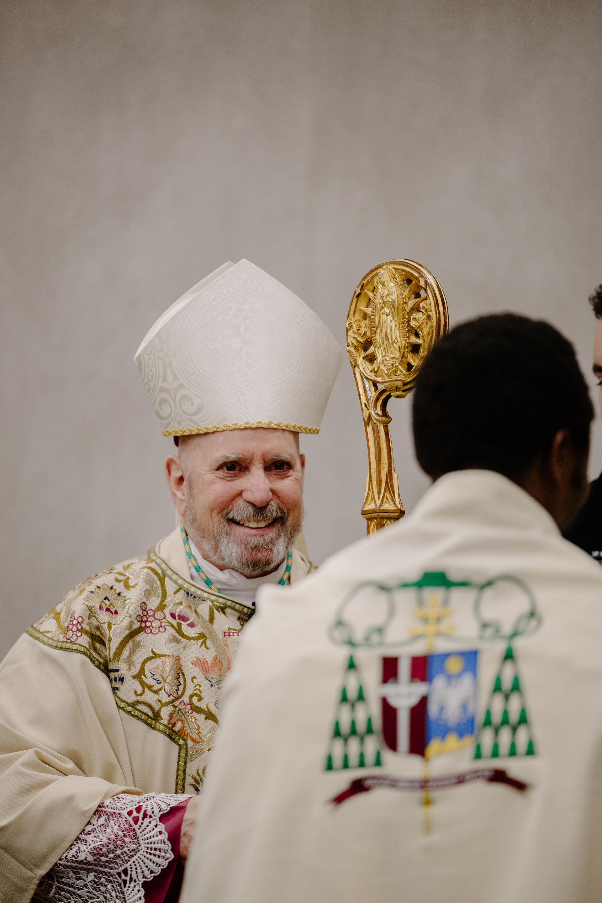 A smiling clergy member in a decorative robe and miter holds an ornate staff. Another person in a robe with crest faces away. Neutral backdrop.