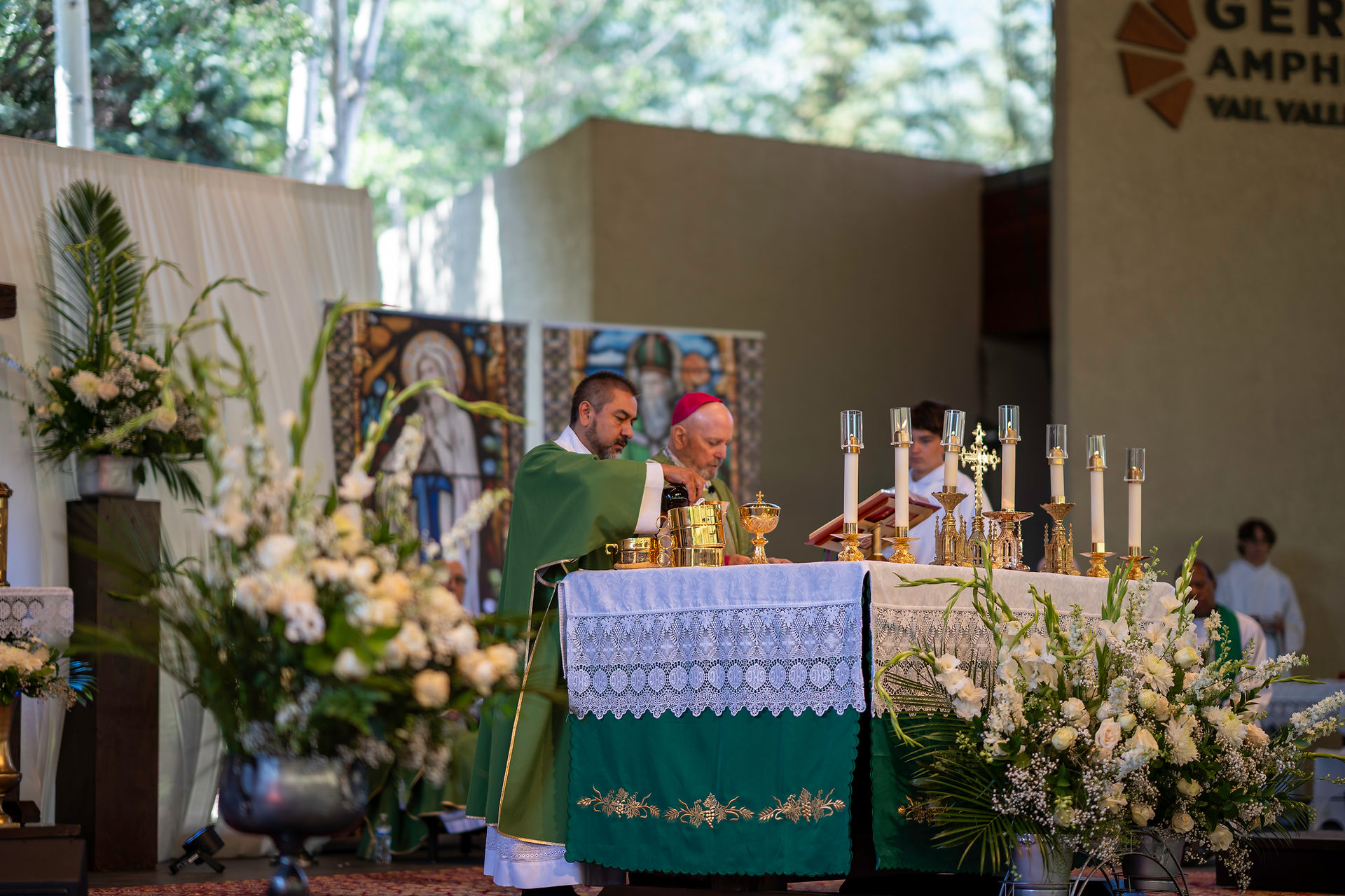 Clergy in green vestments lead a religious ceremony at an altar adorned with candles and flowers. A tapestry and text in the background.
