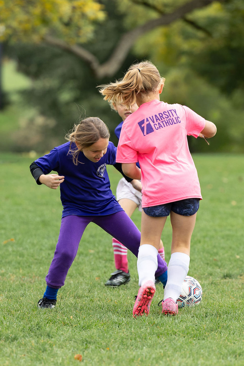 Children playing soccer on a grassy field, one in a pink VARSITY CATHOLIC shirt battling for the ball, trees blurred behind.