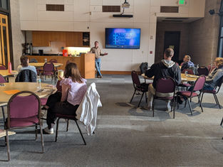 A speaker presents in a conference room with round tables. Attendees listen attentively. Screen displays "VIRTUS: Excellence Builds Trust."