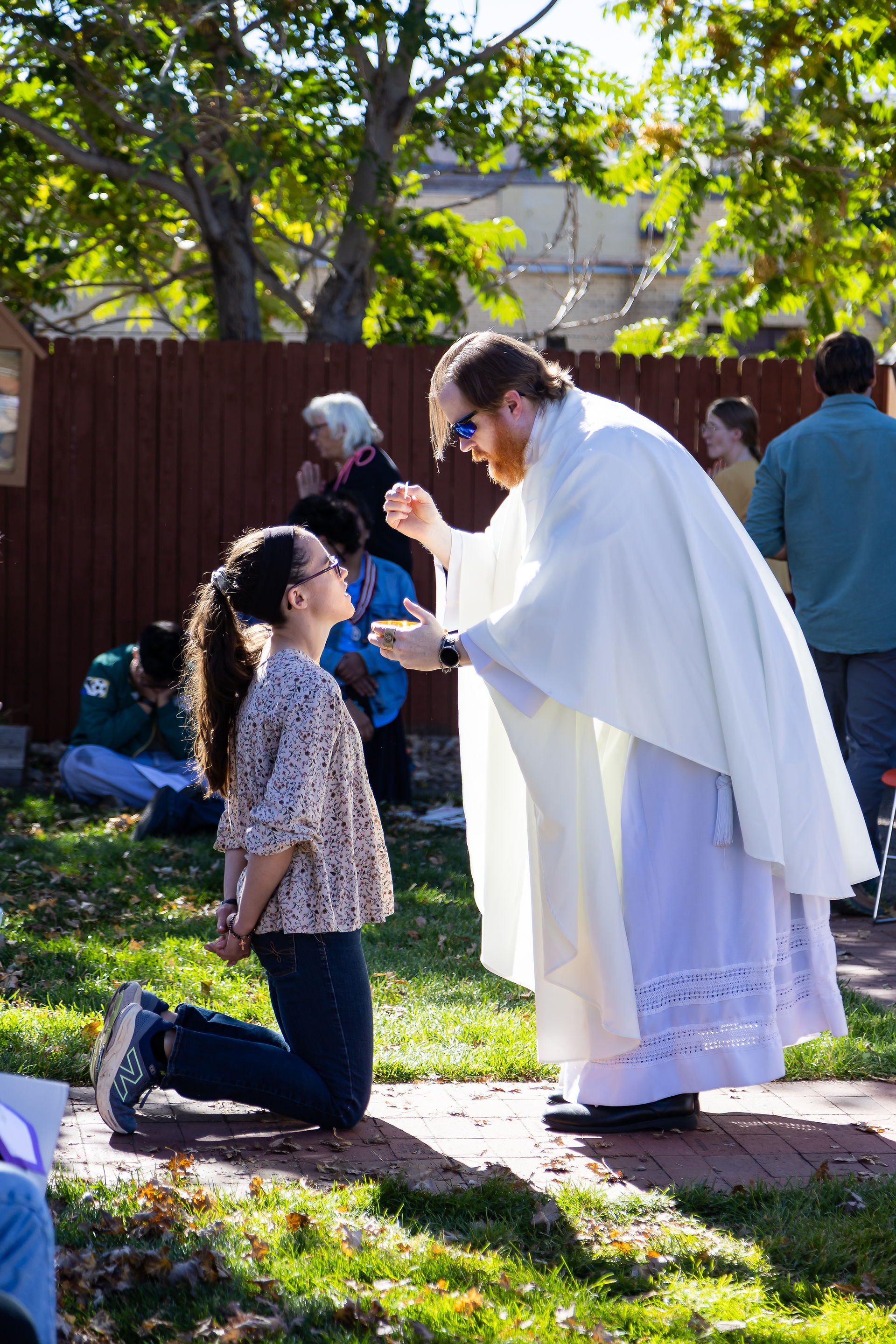 A person in a white robe gives the Eucharist to a kneeling woman outdoors. Others in background, green grass, trees, and sunlight create a serene setting.