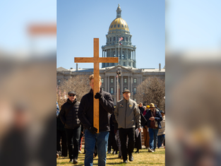 A man carries a large wooden cross in front of a crowd by a domed building. Clear sky and flags in the background. The mood is solemn.