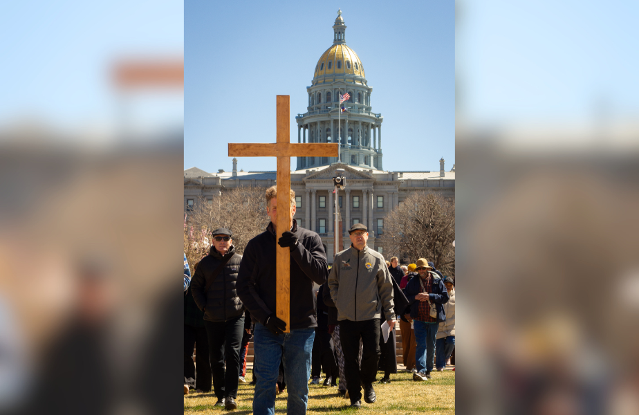 A man carries a large wooden cross in front of a crowd by a domed building. Clear sky and flags in the background. The mood is solemn.