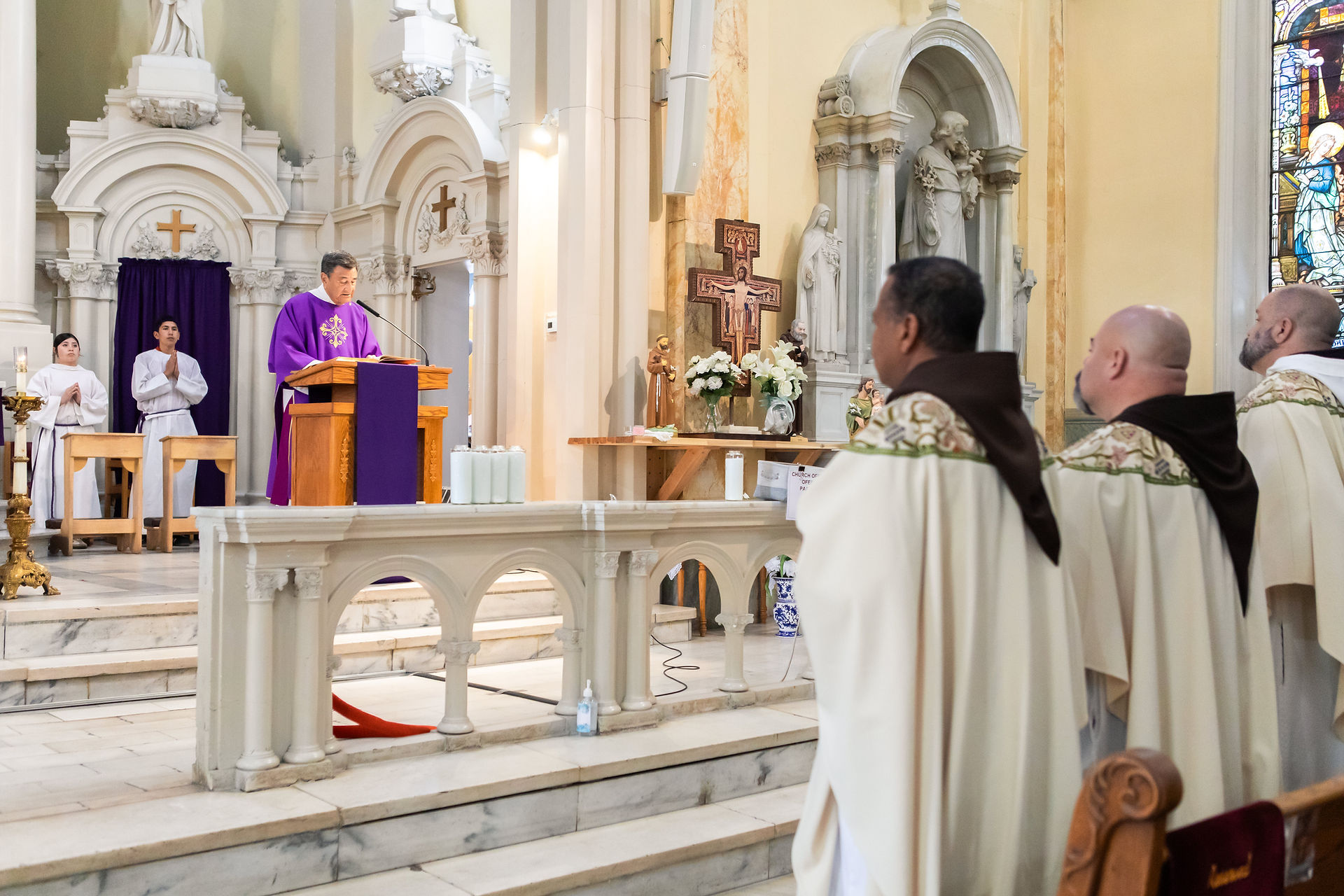 A deacon in purple robes speaks at a church altar. Clergy in white robes and congregation listen. Stained glass, religious statues visible.
