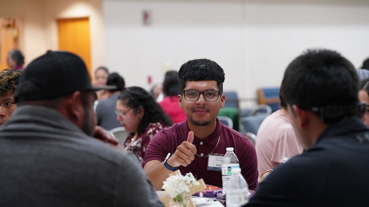 Man in glasses and maroon shirt gives thumbs-up at a table with others in a casual setting. Water bottle and flowers are on table.