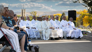Clergy in white robes sit on folding chairs outdoors. They hold papers, surrounded by seated attendees. Blue sky and autumn trees in background.