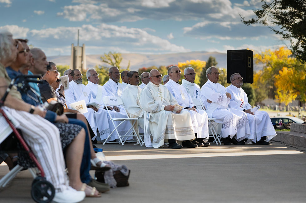 Clergy in white robes sit on folding chairs outdoors. They hold papers, surrounded by seated attendees. Blue sky and autumn trees in background.