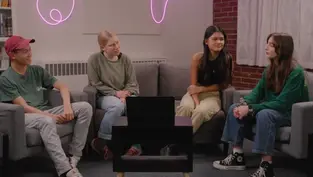 Four people sit on chairs around a table with a laptop in a cozy room. A pink neon light decorates the brick wall, creating a relaxed vibe.