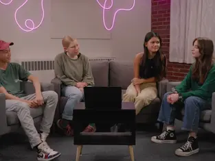 Four people sit on chairs around a table with a laptop in a cozy room. A pink neon light decorates the brick wall, creating a relaxed vibe.