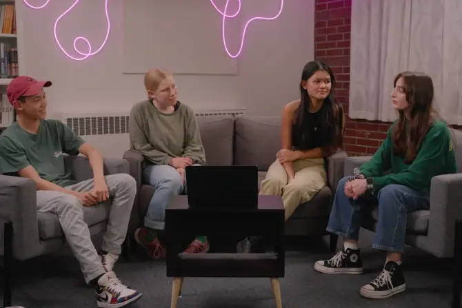 Four people sit on chairs around a table with a laptop in a cozy room. A pink neon light decorates the brick wall, creating a relaxed vibe.