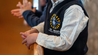 Child in a St. Mary Catholic Virtue School vest claps, hands resting on a wooden rail. Blurred background creates a focused, warm atmosphere.