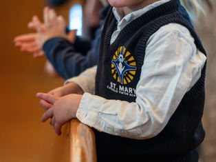 Child in a St. Mary Catholic Virtue School vest claps, hands resting on a wooden rail. Blurred background creates a focused, warm atmosphere.
