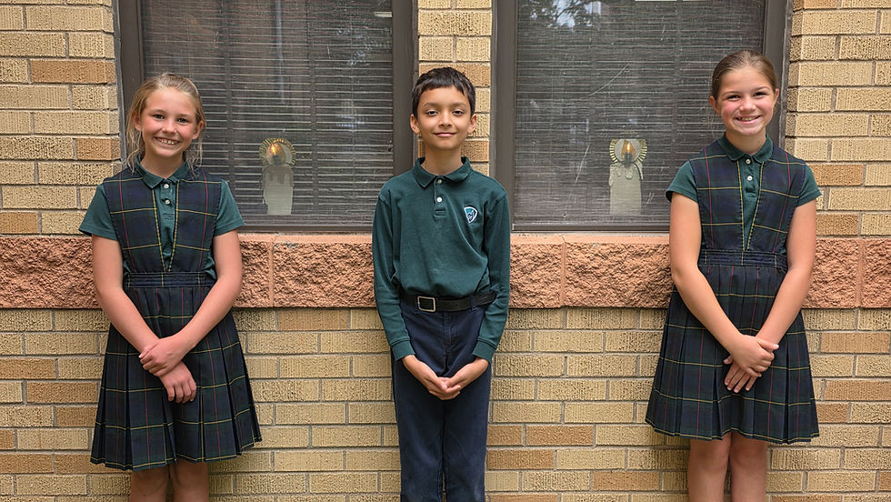Three smiling schoolchildren in green uniforms stand against a tan brick wall outside.
