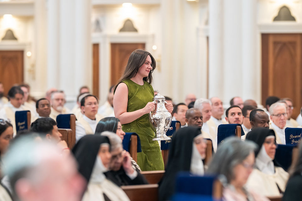 Woman in green dress carries silver vessel in a crowded church with priests and nuns. Blue seat covers say "Reserved Priests."