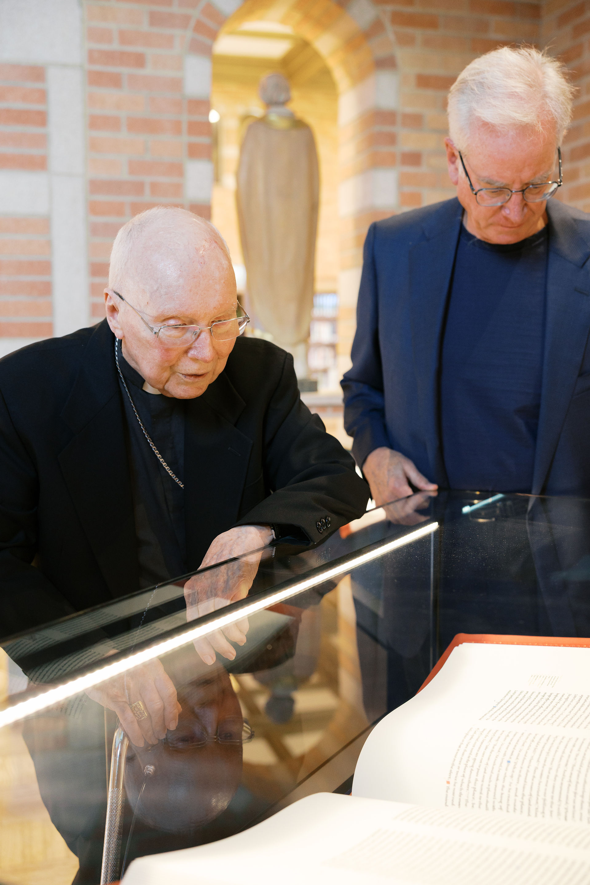 Two men in a library, examining a book through a glass case. One wears black, the other blue; brick wall background; focused.