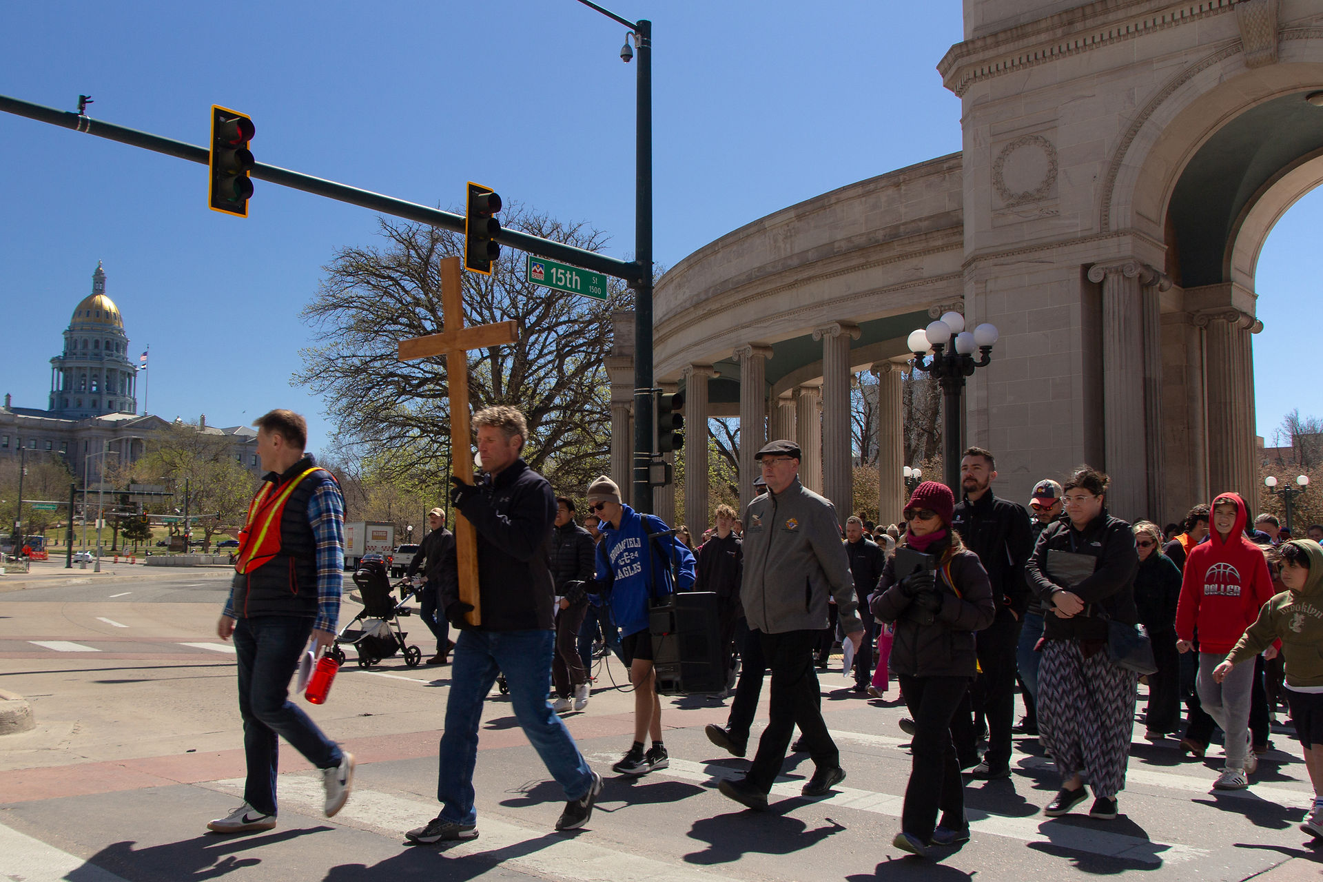 A group of people walk together across a city intersection, led by a man carrying a wooden cross during a public prayer procession.