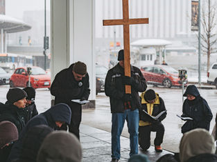 People kneel and read in a snowy urban setting. One holds a large wooden cross, creating a solemn and reflective atmosphere.