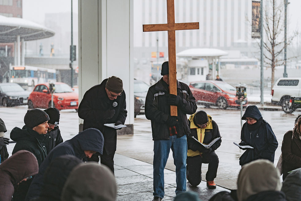 People kneel and read in a snowy urban setting. One holds a large wooden cross, creating a solemn and reflective atmosphere.