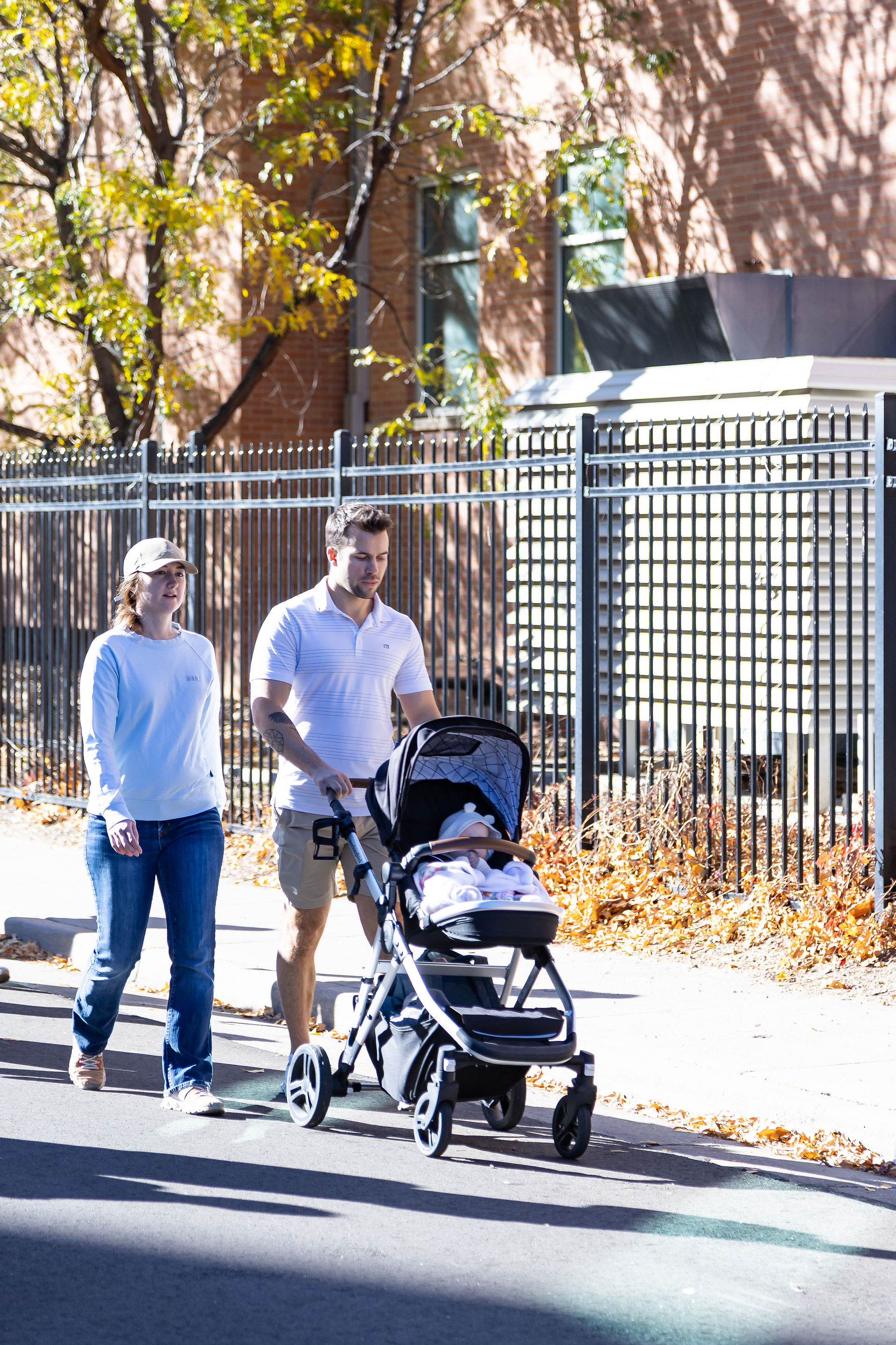 Couple walking with a stroller on a sunny sidewalk. Autumn leaves on the ground, brick building and black fence in the background. Casual mood.