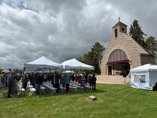 Outdoor ceremony with people gathered under white tents near a stone chapel. Cloudy sky, green grass, solemn mood.