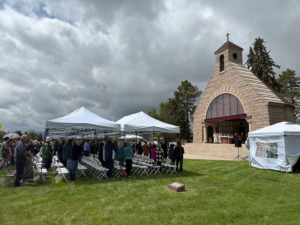 Outdoor ceremony with people gathered under white tents near a stone chapel. Cloudy sky, green grass, solemn mood.