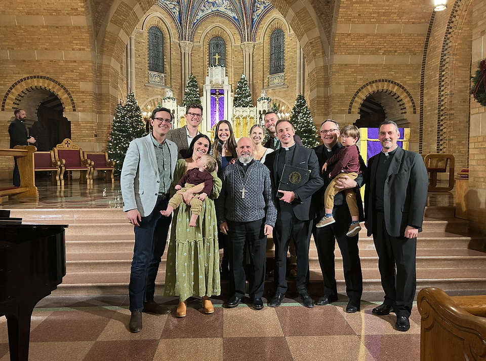 A group of people smiling in a church with Christmas trees. A person holds a child, another holds a plaque. Stone arches and altar in the background.