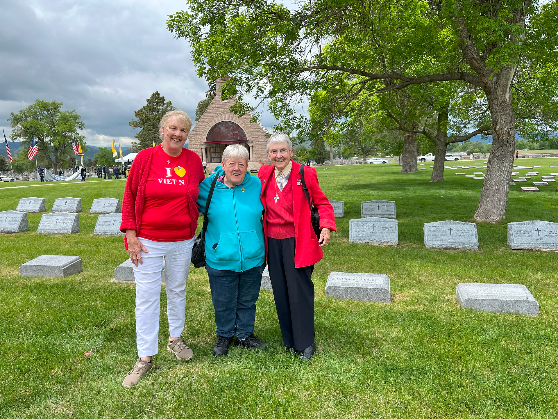 Three older women stand in a cemetery, in front of headstones and a tree. A brick outdoor chapel can be seen in the distance. It's a cloudy day.