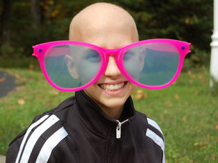 Child smiling, wearing oversized pink glasses outdoors on a grassy background. Dressed in a black and white jacket, exuding joy.