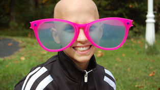 Child smiling, wearing oversized pink glasses outdoors on a grassy background. Dressed in a black and white jacket, exuding joy.