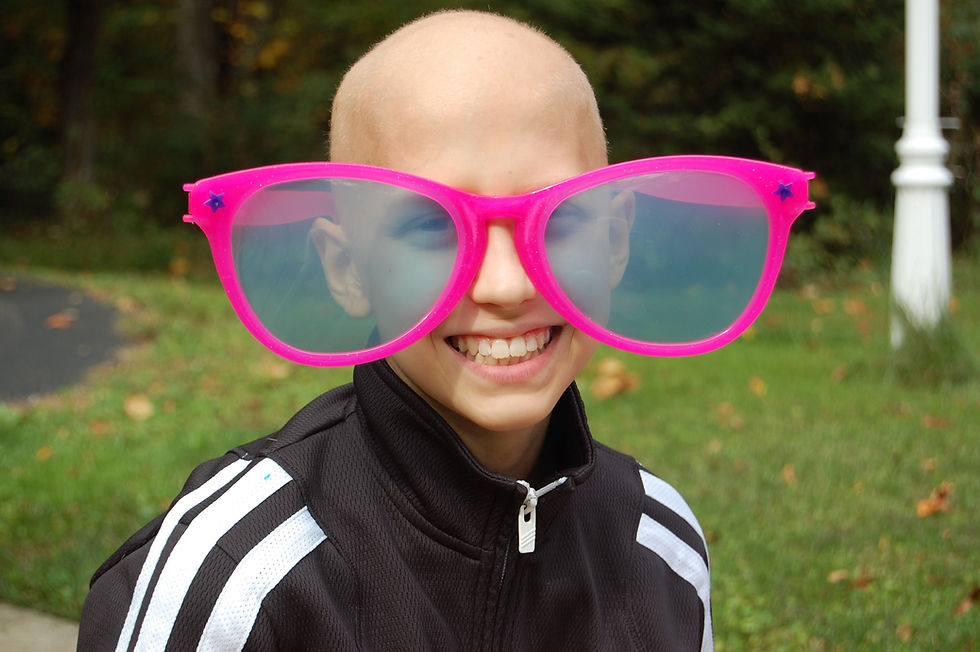 Child smiling, wearing oversized pink glasses outdoors on a grassy background. Dressed in a black and white jacket, exuding joy.