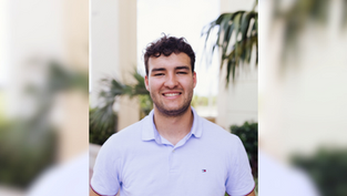 Smiling man in a light blue shirt stands outside with palm trees in the background. Bright day, relaxed mood.
