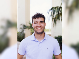 Smiling man in a light blue shirt stands outside with palm trees in the background. Bright day, relaxed mood.