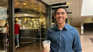 Smiling man in a blue shirt holds iced coffee outside a café with "More Coffee" sign. Warm lighting and wooden floors in the background.