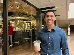 Smiling man in a blue shirt holds iced coffee outside a café with "More Coffee" sign. Warm lighting and wooden floors in the background.