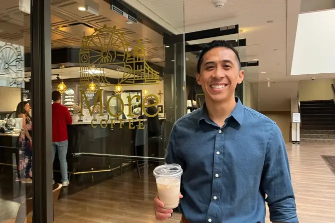 Smiling man in a blue shirt holds iced coffee outside a café with "More Coffee" sign. Warm lighting and wooden floors in the background.