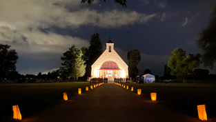 Illuminated path leading to a lit chapel at night, with glowing lanterns and cloudy sky. Trees frame the serene, inviting scene.