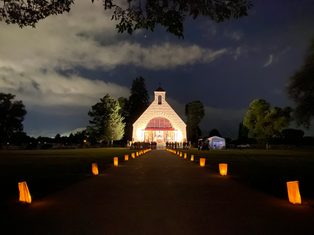 Illuminated path leading to a lit chapel at night, with glowing lanterns and cloudy sky. Trees frame the serene, inviting scene.