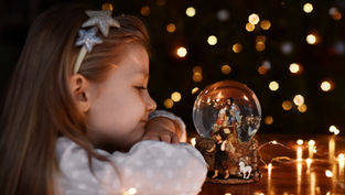 Girl with star headband gazes at a nativity snow globe on a table, surrounded by warm fairy lights. Bokeh effects create a cozy atmosphere.