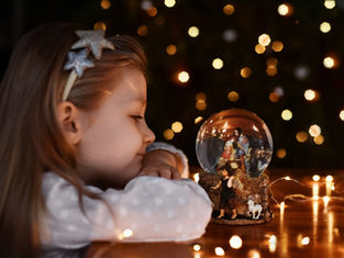 Girl with star headband gazes at a nativity snow globe on a table, surrounded by warm fairy lights. Bokeh effects create a cozy atmosphere.