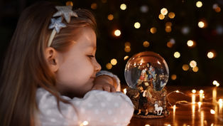 Girl with star headband gazes at a nativity snow globe on a table, surrounded by warm fairy lights. Bokeh effects create a cozy atmosphere.