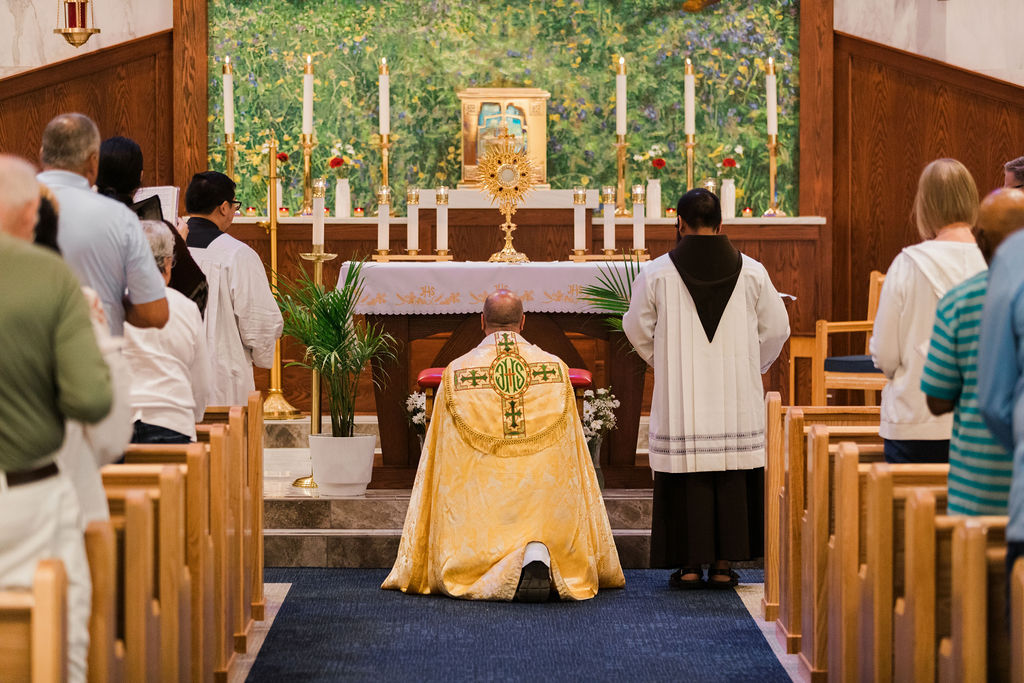 Priest kneels before altar at church service, facing golden monstrance and candles, while congregation stands in pews.