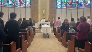 People in a chapel attend a service led by a priest. A casket draped in white is in the center. Stained-glass windows in the background.