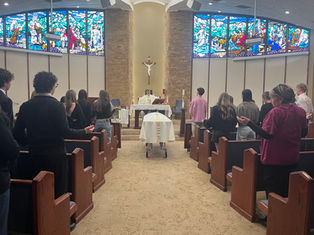 People in a chapel attend a service led by a priest. A casket draped in white is in the center. Stained-glass windows in the background.