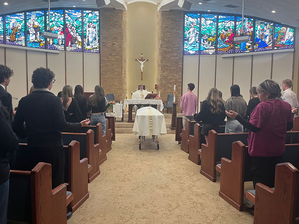 People in a chapel attend a service led by a priest. A casket draped in white is in the center. Stained-glass windows in the background.