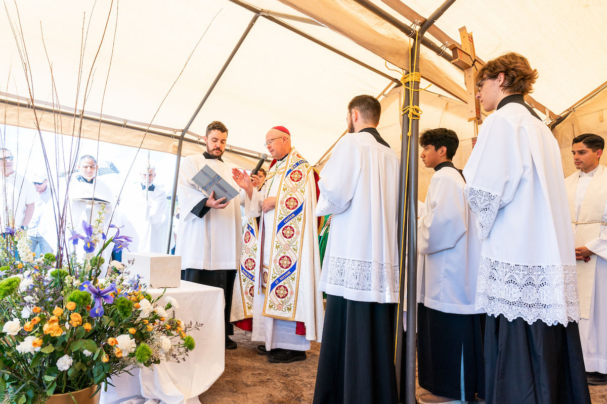 Bishop speaks with microphone to altar servers in white robes under a tent, beside a flower-filled altar in a solemn ceremony
