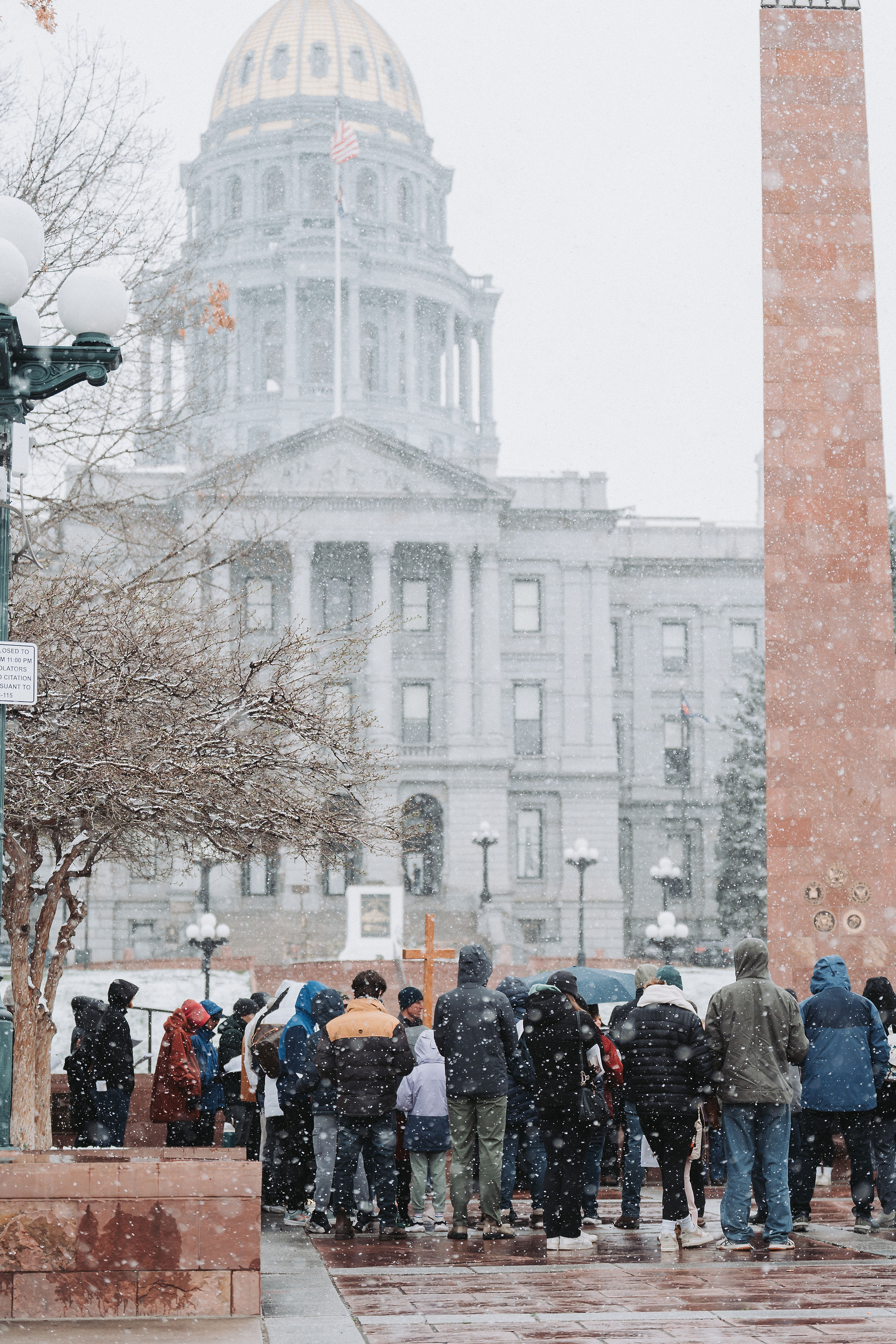 A group of people stand in a snowy scene outside a grand building with a dome. They wear winter clothing, suggesting a cold, solemn gathering.