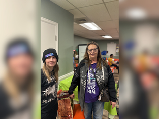 Two women in casual attire stand smiling in a room with lime-green tablecloths. One wears a beanie, the other a jacket over a purple shirt.