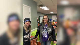 Two women in casual attire stand smiling in a room with lime-green tablecloths. One wears a beanie, the other a jacket over a purple shirt.